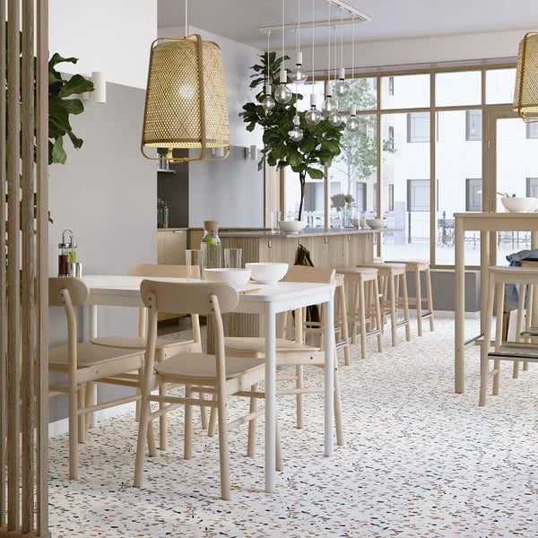 A bright and modern café interior with large floor-to-ceiling windows that let in natural light. The space features white terrazzo flooring with colorful speckles and light wood furniture, including simple square tables, matching chairs, and tall bar stools. Overhead, there are a mix of pendant lights—some with woven rattan shades and others with exposed glass bulbs. Several large green plants add freshness to the room, while glassware and minimalist table settings contribute to the clean, Scandinavian-inspired aesthetic. The overall atmosphere is airy, warm, and welcoming.