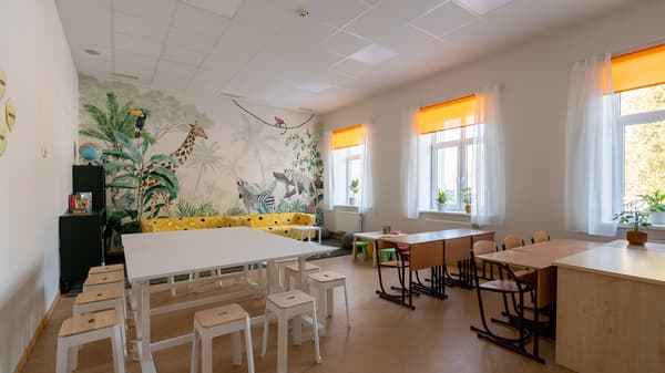 A bright and multifunctional after-school space with study tables, lounge areas, and a tropical jungle mural featuring wild animals. Yellow sofas, wooden furniture, and large windows with orange blinds create a cheerful and welcoming environment for studying, socialising, and relaxation.