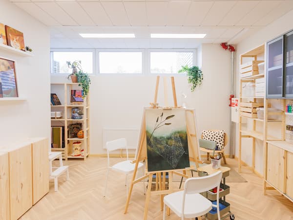 A bright and organised art studio featuring wooden shelves, a central easel with a painting, chairs, and neatly arranged art supplies.