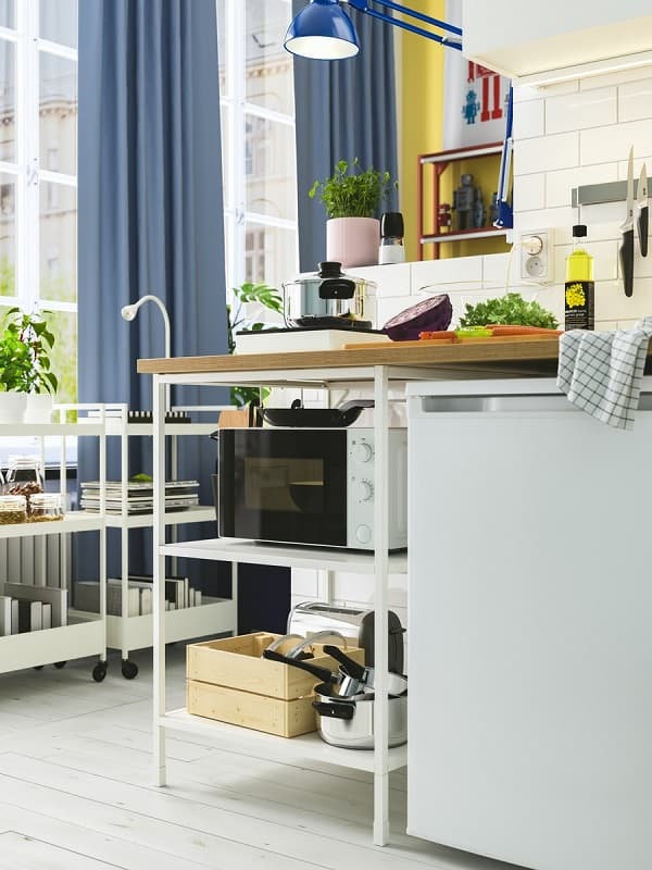 A bright and small kitchen area with white ENHET kitchen combination, portable induction hob and a small fridge. White trolleys store cookbooks and plants.