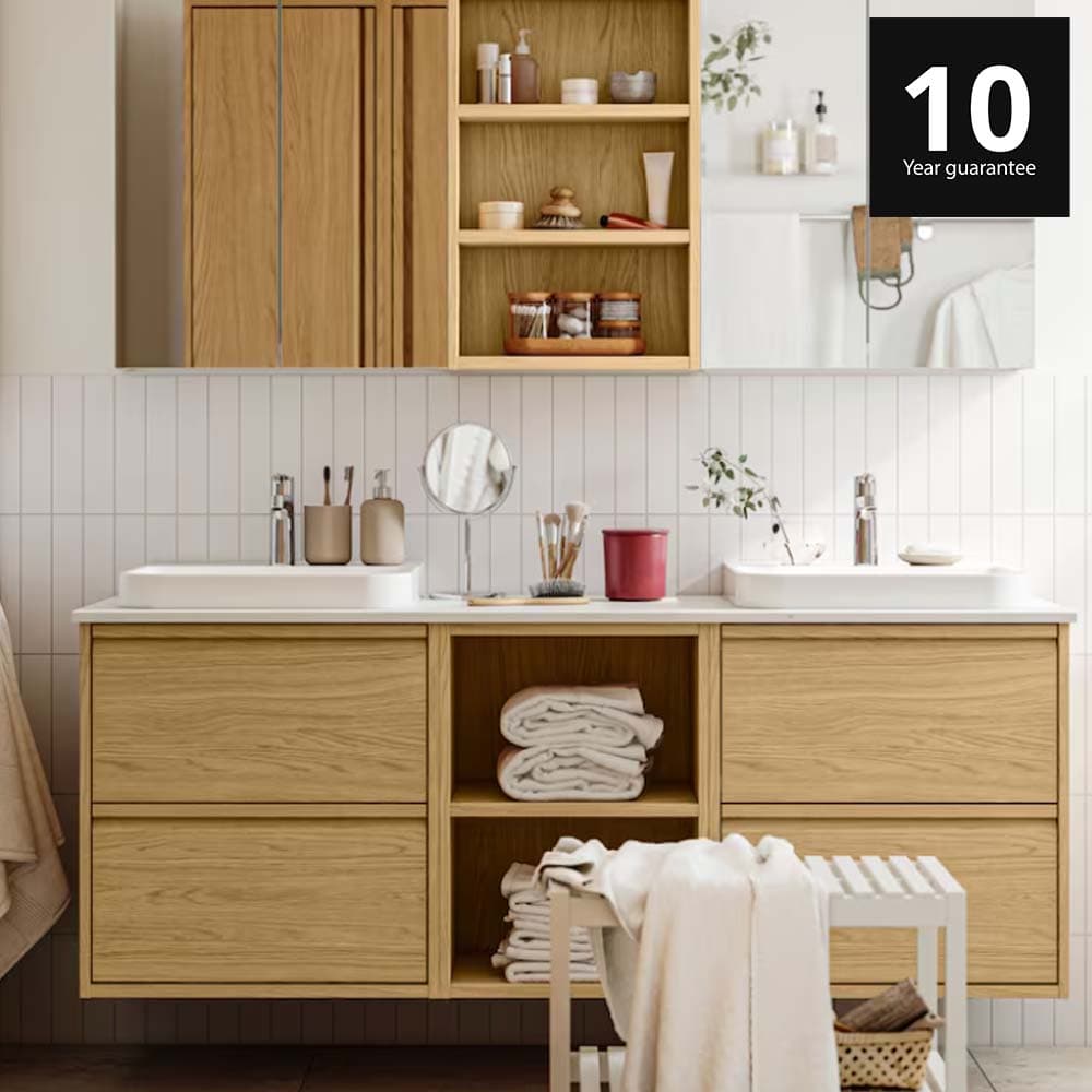 A bright bathroom has ÄNGSJÖN bathroom furniture in oak effect, including wall cabinets and a wash-stand with drawers. 10-year guarantee logo on top right corner.