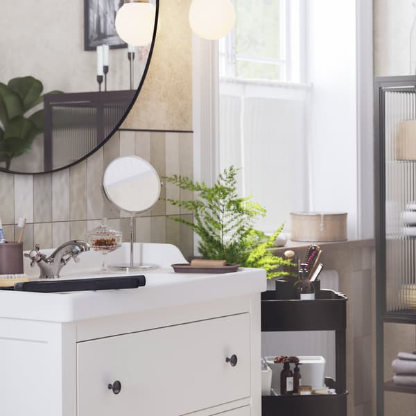 A bright bathroom with a window has a white HEMNES washstand with a round mirror above it and a black trolley next to it.