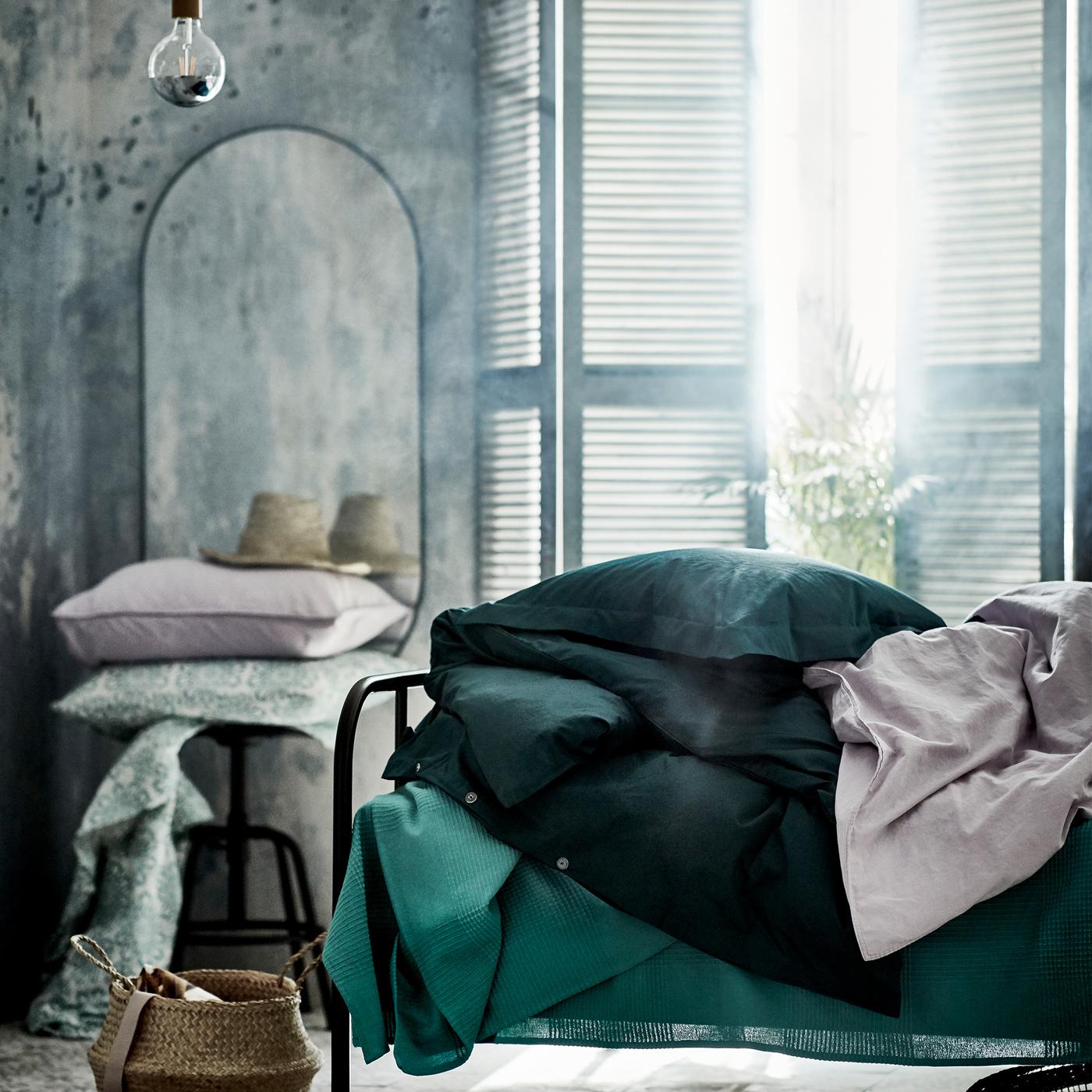 A bright bedroom with a bed made with green bed linen. Beside is a mirror and a stool holding extra pillows.