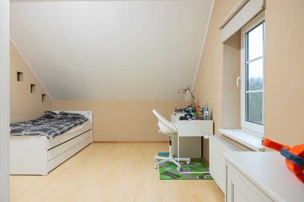 A bright children’s room under a sloped ceiling with a bed along the wall, light wood flooring, and toys neatly arranged near the window.