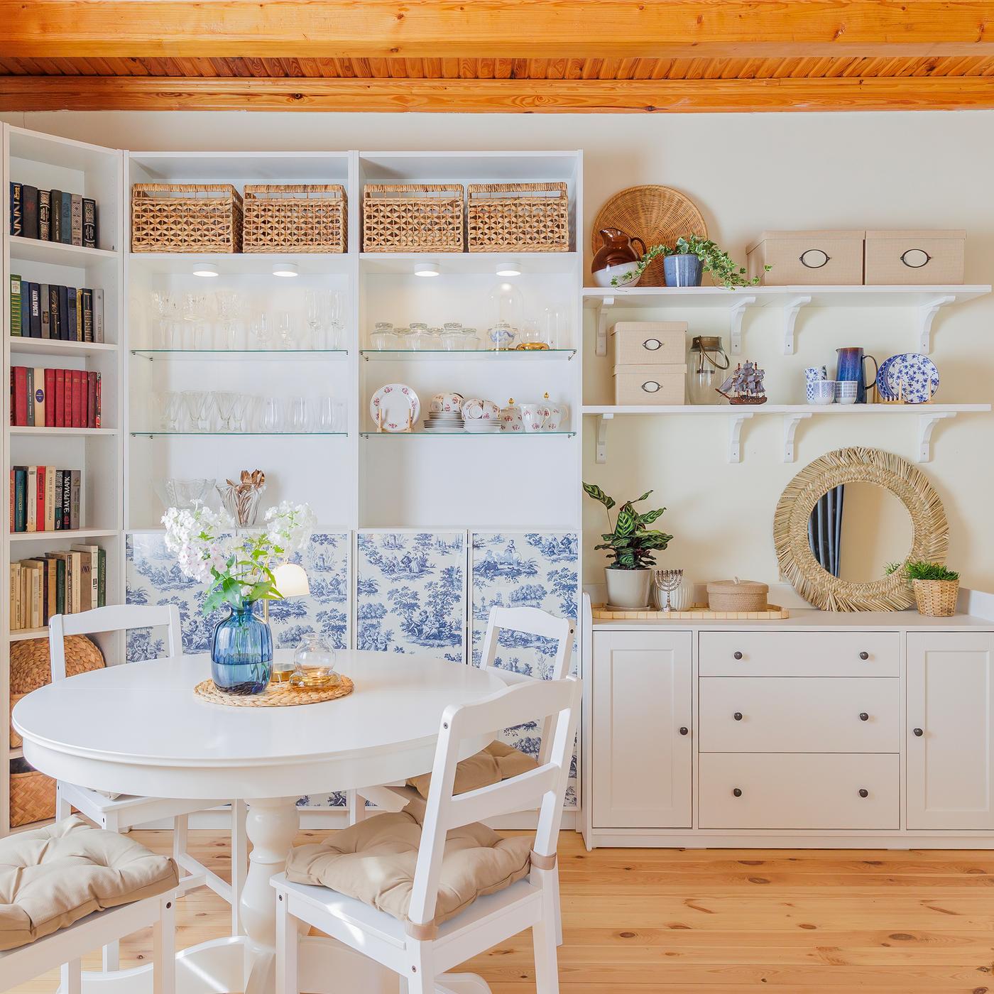 A bright dining area featuring a round dining table with white chairs, surrounded by a bookcase filled with books. Soft blue accents throughout the room add a calm and inviting atmosphere.