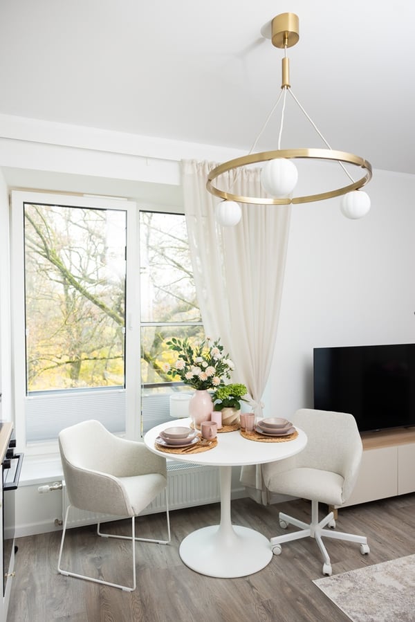A bright dining area with a round table, chairs, a modern ceiling light, and a large window.