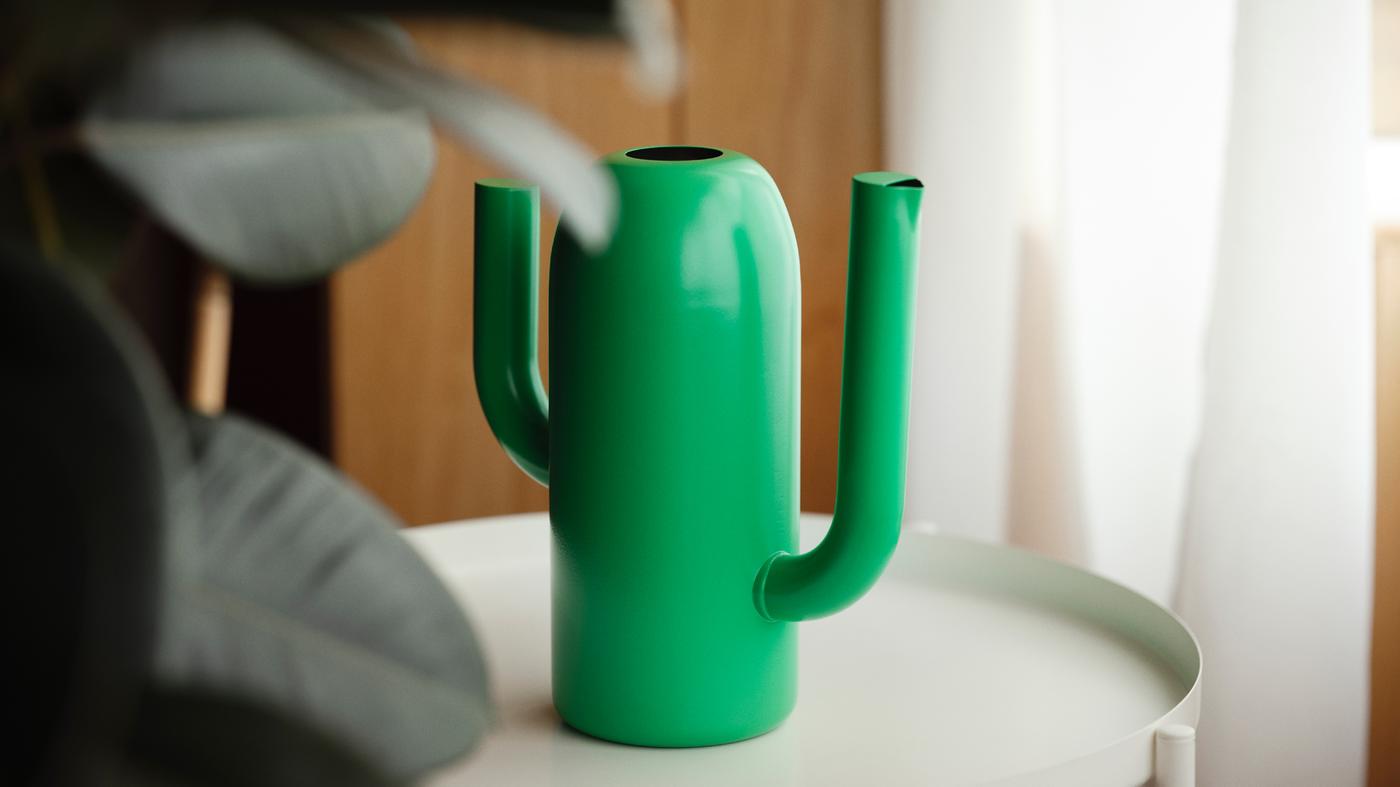 A bright green ÄRTBUSKE vase and watering can with two cactus-like arms sits on a table with a plant in front of it.