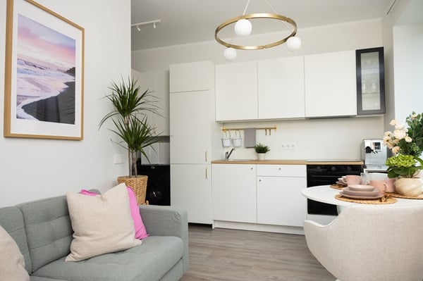 A bright living room with an open-plan kitchen, white cabinets, a round dining table, plants, and a modern ceiling light.