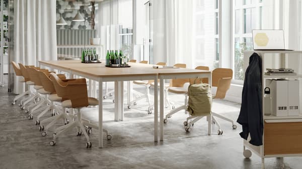 A bright meeting room with tables and chairs in light wood and white tones, sheer white curtains and a glass wall.