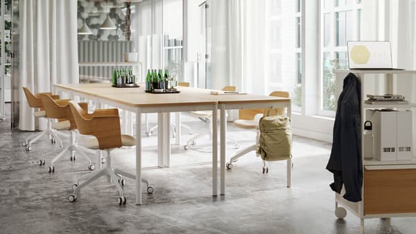 A bright meeting room with tables and chairs in light wood and white tones, sheer white curtains and a glass wall.