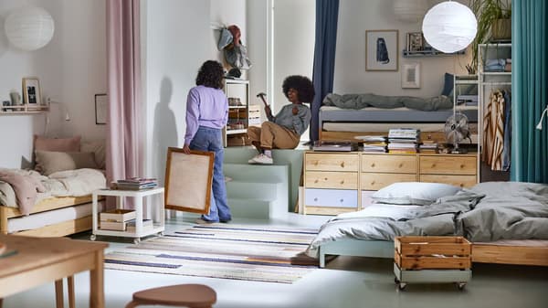 A bright room with two women hanging a picture, a bed, white pendants above and a rug on the floor.