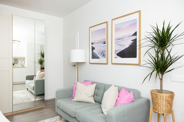 A bright seating area with a light-coloured sofa, decorative cushions, framed artwork on the wall, and a houseplant in the corner.