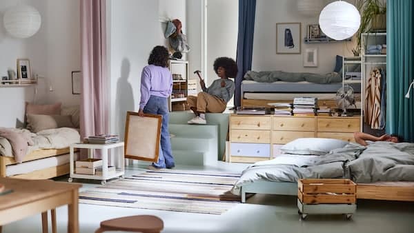 A bright shared room with two women hanging a picture and a man in one of three beds, white pendants above and a rug on the floor.