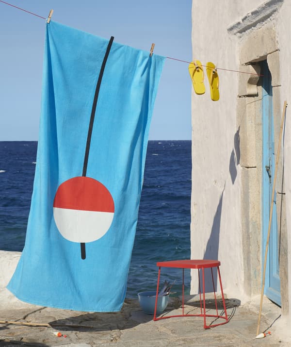A brightly coloured beach towel and flip flops hanging from a washing line next to a red side table by the sea.