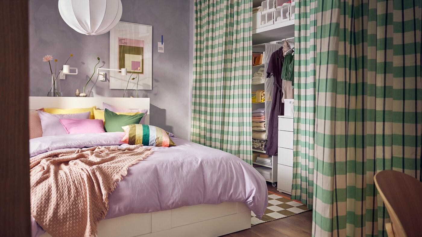 A BRIMNES bed in a lilac room covered in lilac NATTSVÄRMARE bed linen. A green GLANSHAGTORN curtain covers clothing storage.