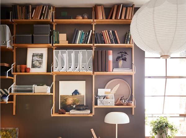 A brown-painted wall with a wall-mounted shelf combination in bamboo with books, decorative items and white magazine files.
