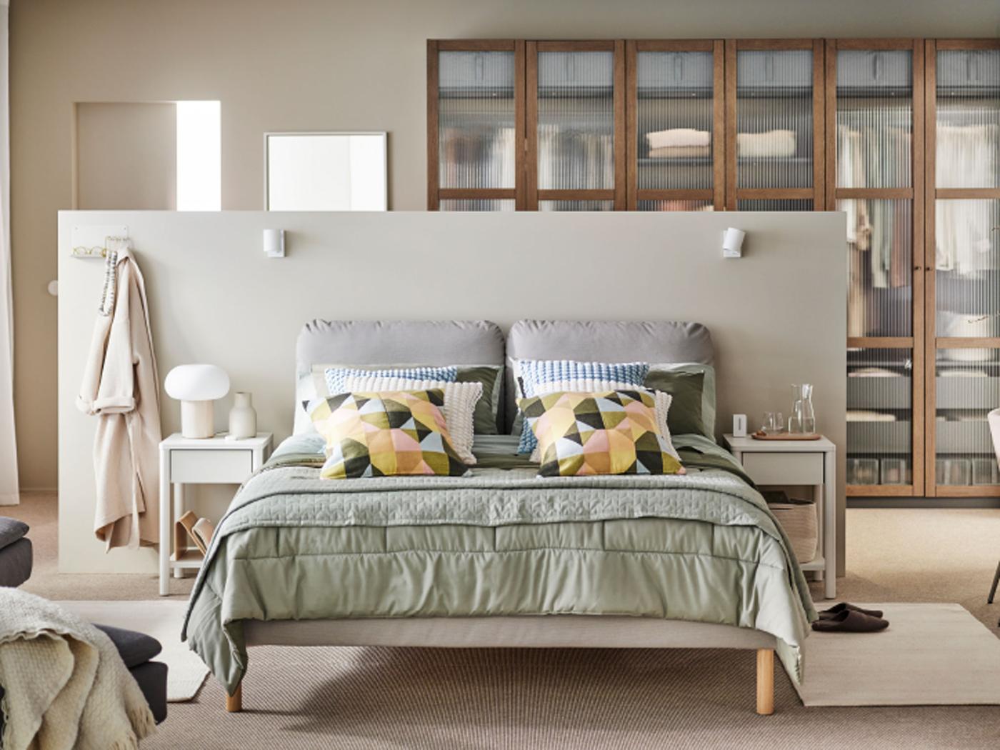 A brown SAGESUND upholstered bed in a beige bedroom with a grey-green KÖLAX bedspread and cushions in SVARTHÖ covers on top.
