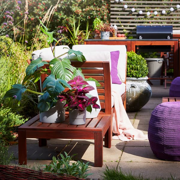 A brown-stained table with plants in grey plant pots and a white table lamp. A purple pouffe stands beside on the ground.