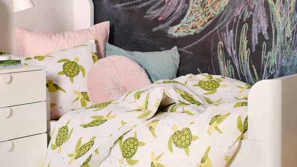 A BUSUNGE white bed is dressed in a BLÅVINGAD green turtle duvet cover with cushions in front of a chalkboard wall.