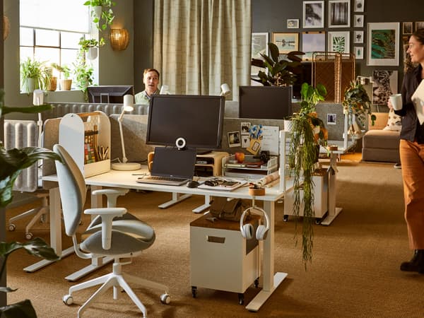 A busy office in earthy colours, natural materials, plants, and a large picture wall. Two colleagues are having a conversation.