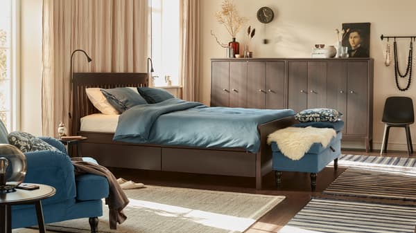 A calm bedroom with beige walls, linen curtains, wooden bed, dark blue textiles, bench, and wooden cabinets by the walls.