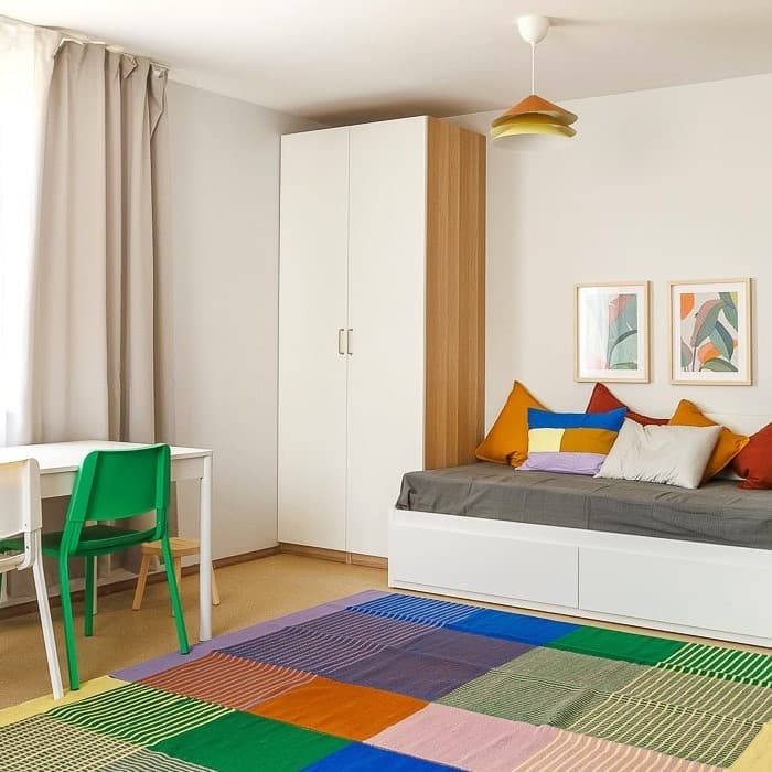 A calm, homely room in a crisis centre featuring a neatly made single bed with cushions, a colourful rug underfoot and a bright green chair tucked beside a small desk.