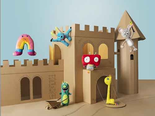 A cardboard castle in front of a blue sky background with several colorful soft toys positioned around the castle.