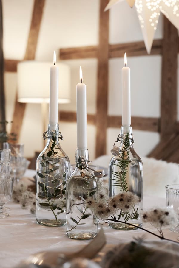 A centrepiece made with candles and fir branches in glass bottles filled with water, displayed on a dining table.