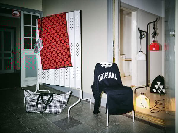 A chair draped with a dark shirt that says ORIGINAL on it, and a white room divider behind it with a red rug draped over it.