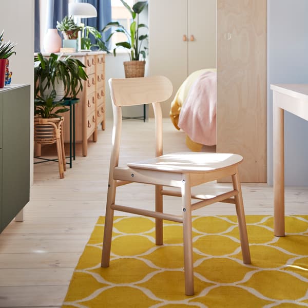A chair in birch stands on a yellow rug. A chest of drawers in birch and green plants stand in a room in the background.