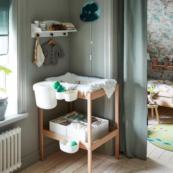 A changing table with accessories in a bedroom. A window on one side, an alcove doubling as a children’s room on the other.