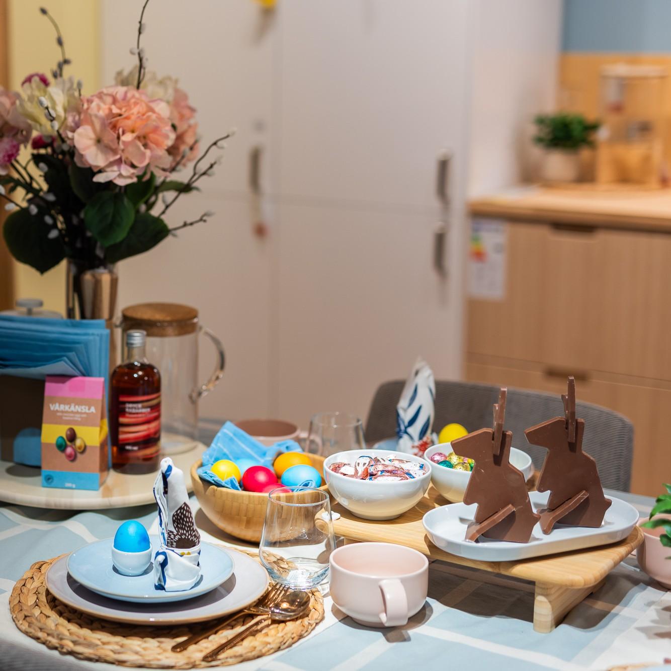A cheerful and vivid Easter table setting in shades of blue and pink. A wooden bowl with colored eggs and a wooden tray with two chocolate bunnies in the center of the table.