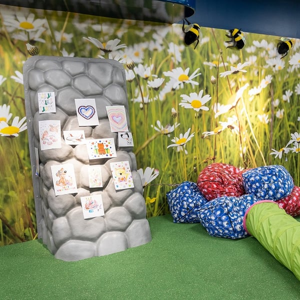 A cheerful children's play area with a daisy-themed wall mural, a stone-like display pillar showcasing colorful kids' drawings, soft red and blue beanbags, and a green crawl tunnel on artificial grass flooring. Plush bees hang playfully from the ceiling above.