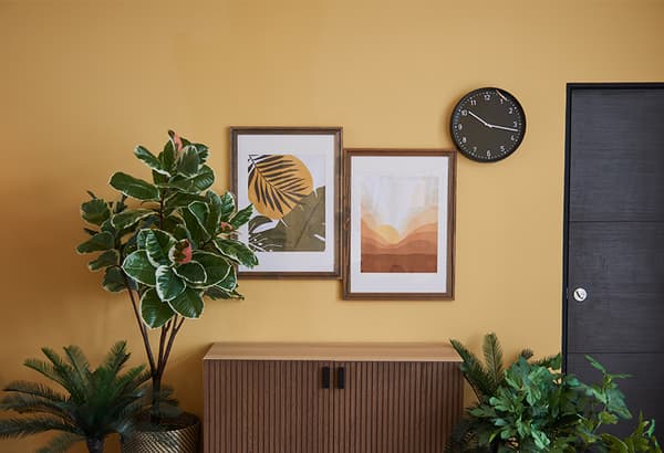 A chest of drawer with frames hung on top and decorative plants by the side.