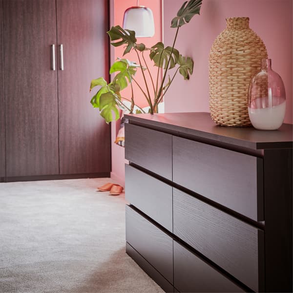 A chest of drawers and a wardrobe in black-brown, a gray rug, a glass vase and a green plant.