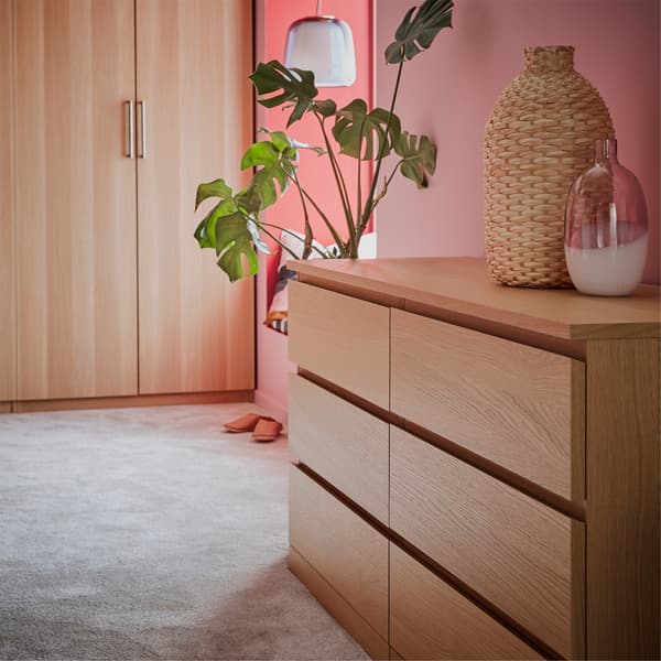 A chest of drawers and a wardrobe in white stained oak effect, a grey rug, a glass vase and a green plant.