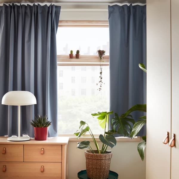 A chest of drawers in birch, a white table lamp, some green plants and blue curtains that cover most of a window.