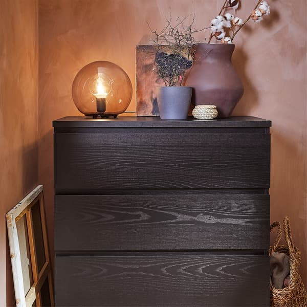 A chest of drawers in black-brown, a terracotta vase, a dark gray plant pot and a gray table lamp.