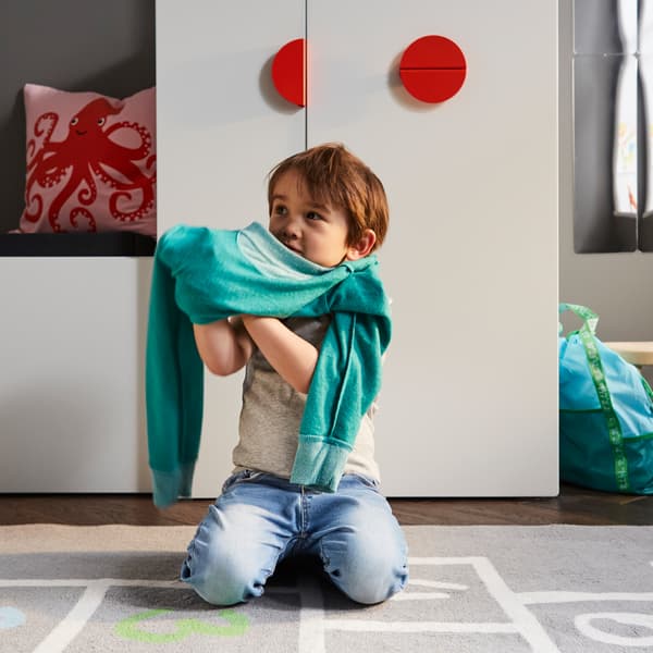 A child getting dressed on a grey rug in front of a white bench with toy storage and a white wardrobe with a pull-out unit.