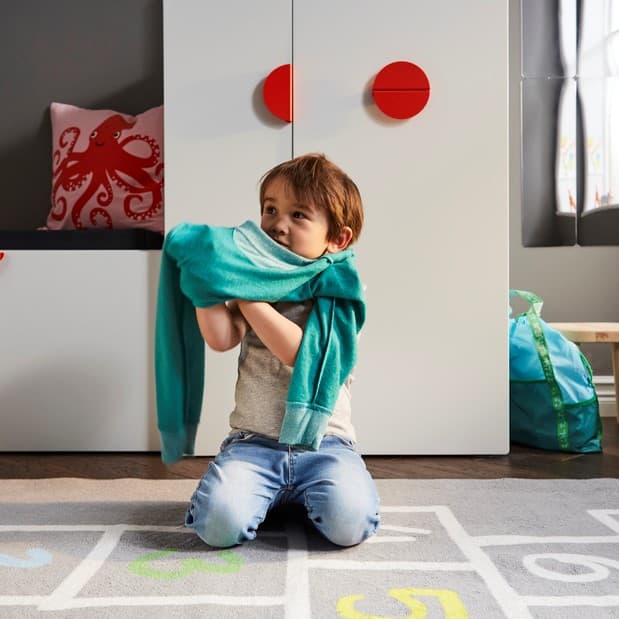A child getting dressed on a grey rug in front of a white bench with toy storage and a white wardrobe with a pull-out unit.