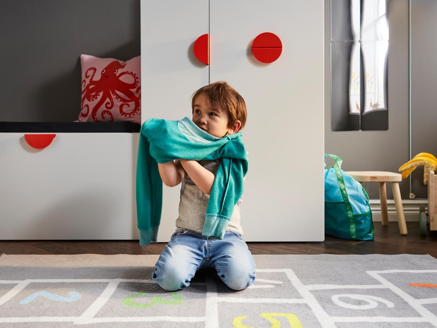 A child getting dressed on a grey rug in front of a white bench with toy storage and a white SMÅSTAD wardrobe with a pull-out unit.