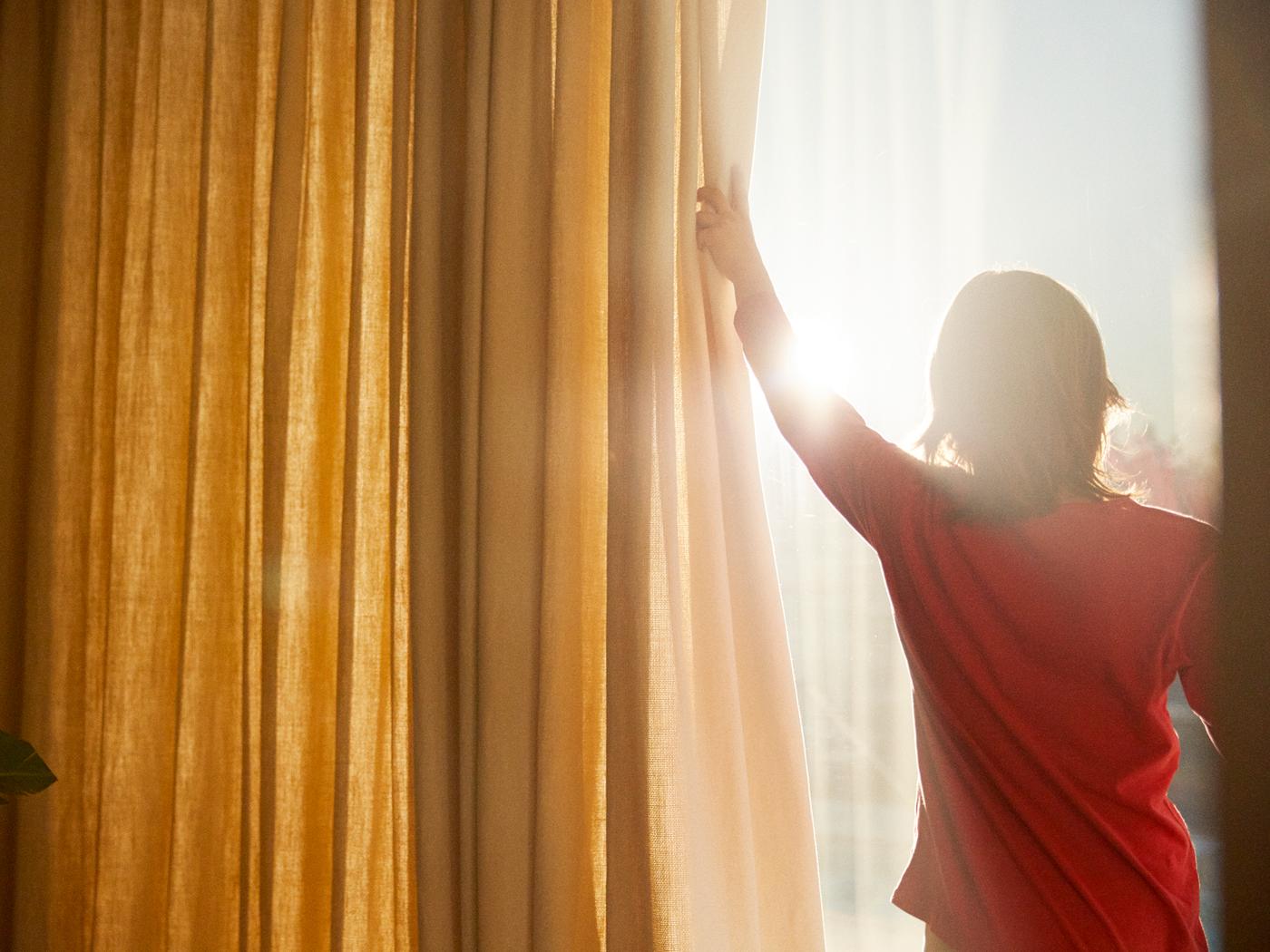 A child in a red t-shirt pulls back yellow and white curtains to reveal the sun blazing through the window.