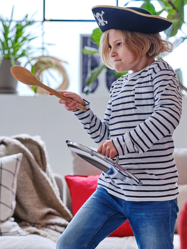 A child in blue jeans, a striped top and a pirate hat plays with a wooden spoon and a saucepan lid in a mock sword fight.