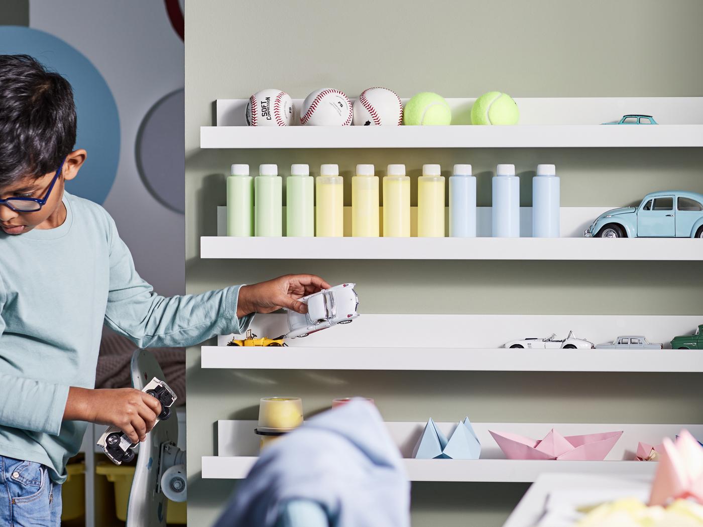 A child playing with toy cars in a bedroom with MOSSLANDA picture ledges on the wall holding toys, paper models and bottles.