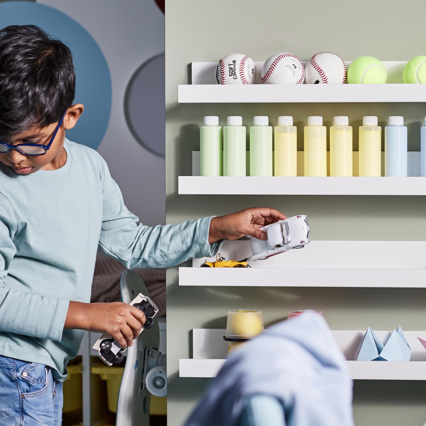 A child putting toy cars on a white picture ledge on a wall with three more picture ledges holding paint bottles and balls.