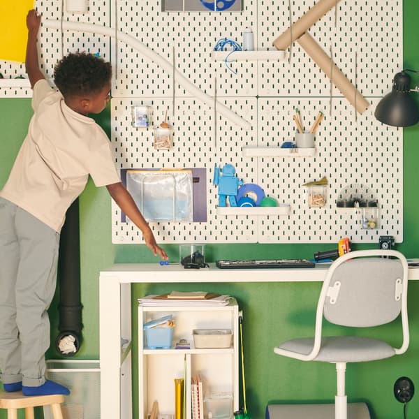 A child reaching across a wall with white pegboards holding various items, above a white desk with a children’s desk chair.