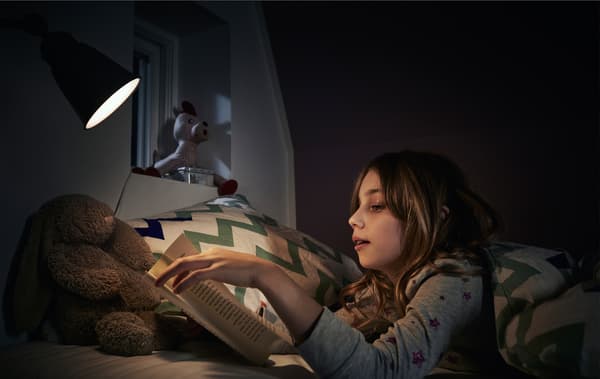 A child reading a book in bed with a reading lamp.