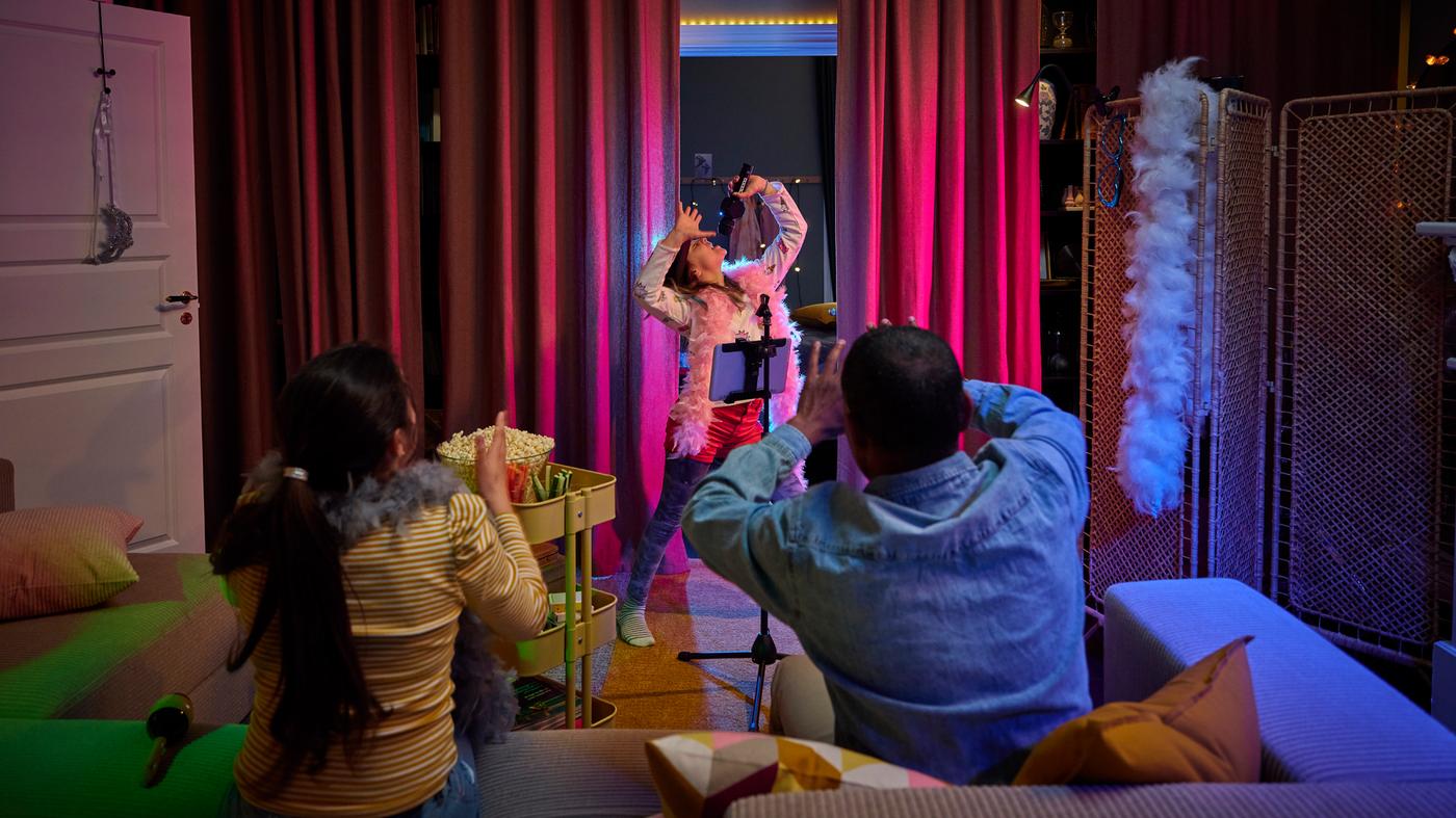 A child singing into a microphone in front of dark red curtains, with two people in a grey/beige JÄTTEBO sofa looking on.