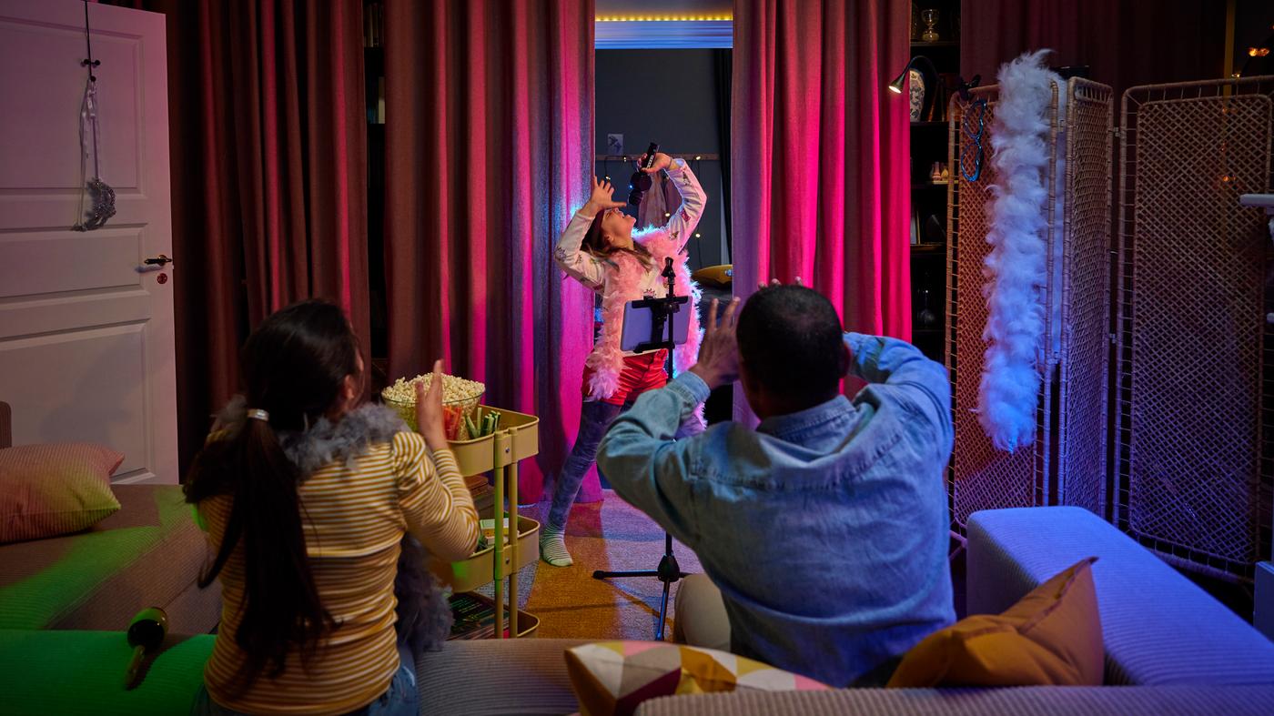 A child singing into a microphone in front of dark red curtains, with two people in a grey/beige JÄTTEBO sofa looking on.