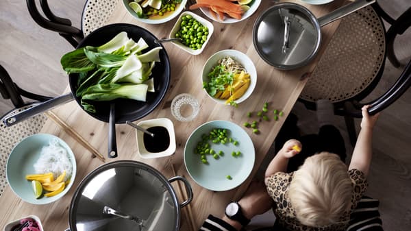 A child sits on an adult’s lap eating finger food at a wooden table with fruit and vegetables in bowls and pans.